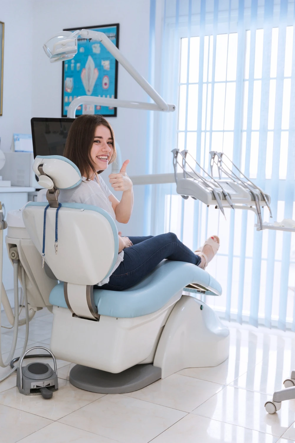 Smiling female patient sitting on chair showing thumb-up in dental clinic<br />
